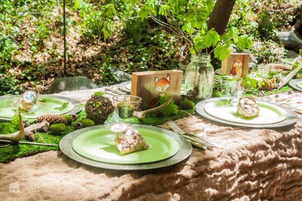 Woodland themed baby shower. Table decorations include, burlap tablecloth, moss tablerunner, pinecones, pods, leaves, logs, moss balls, baby animal images. Place settings are on a wood grain charger with green paper plates.