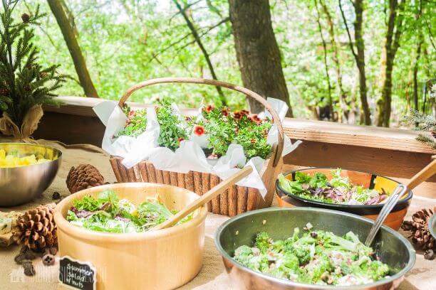 The buffet table is decorated with burlap, pine boughs, pinecones, logs and twigs. The food includes a variety of salads, berries, edible moss cupcakes, and acorn cake pops.