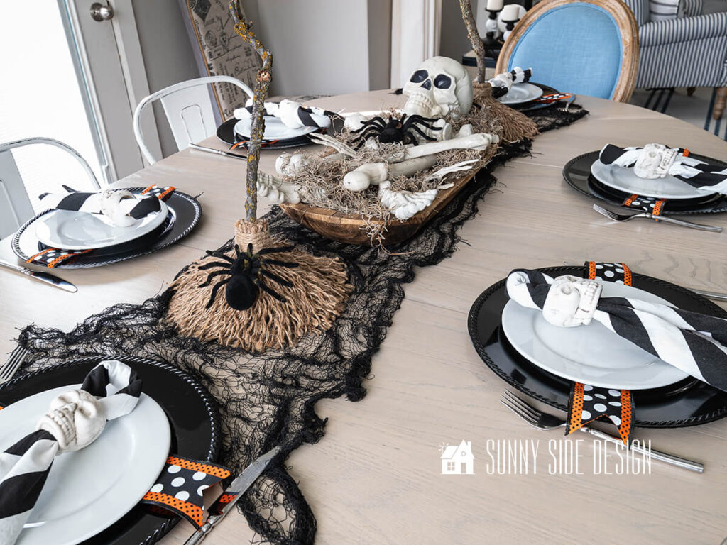The dining table is decorated with a black creepy cloth table runner, dough bowl filled with spanish moss, skeleton bones and black birds. On each side of the dough bowl is a DIY witch broom.