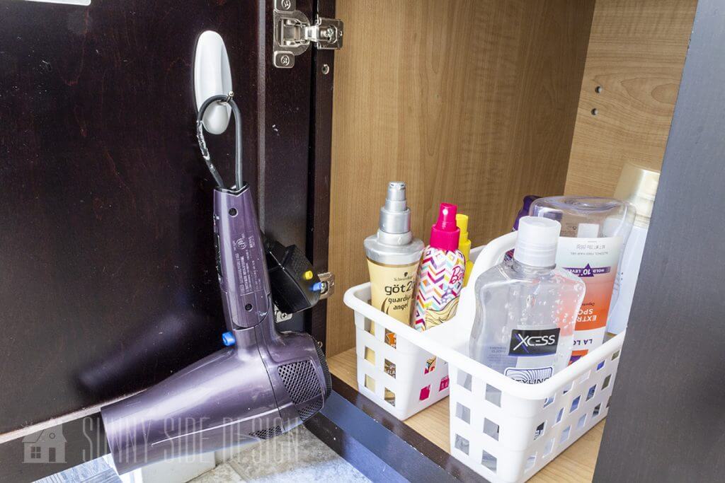 A hair dryer is hung from a command hook on the inside of the bathroom cabinet door. A plastic shower caddy is used to store hair care products.