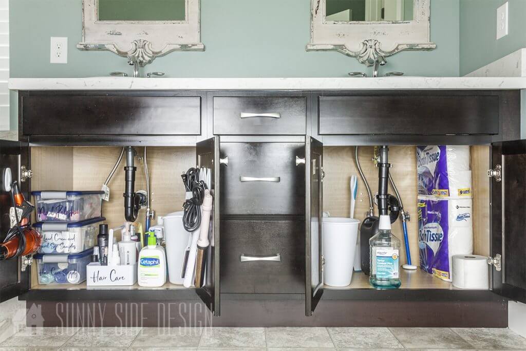 Both sides of the bathroom vanity cabinet are organized by implementing label plastic storage contains as well as hanging hair tools on hook.