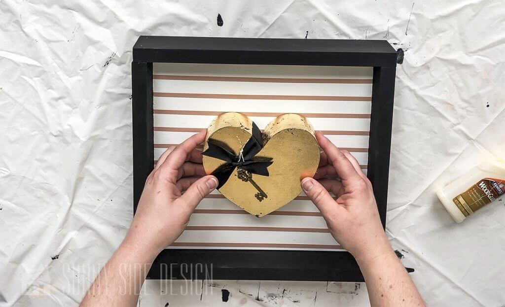 Woman's hands placind gold leaf heart on wood frame.