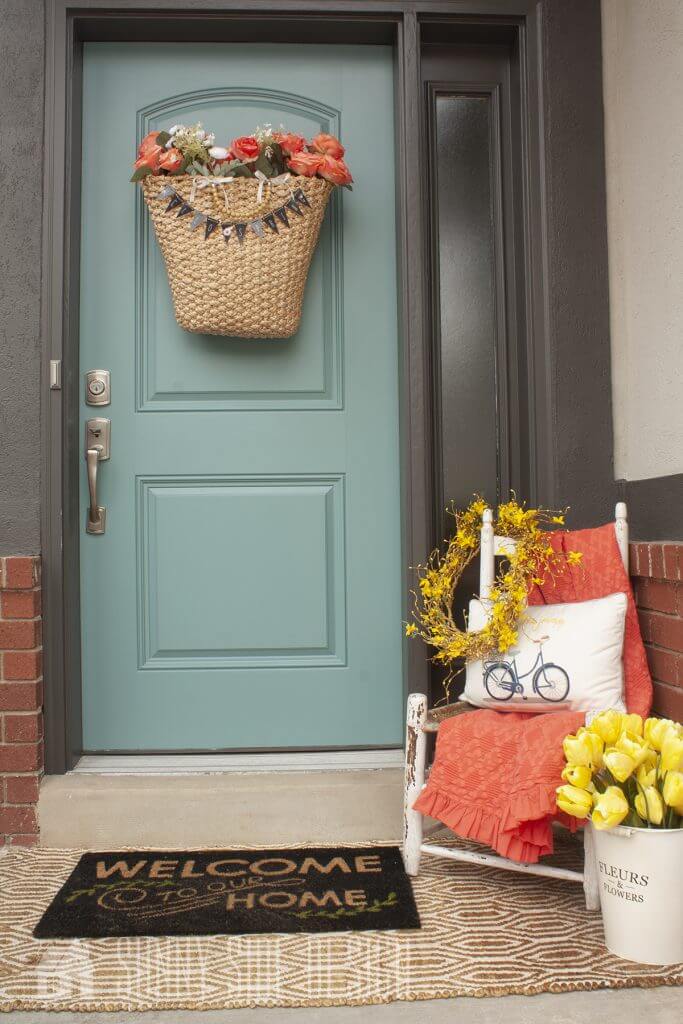 Basket full of flowers with a small spring banner printable "hello spring" on a blue door. Front porch decorated with chair, coral throw, pillow, forsythia wreath and a bucket of tulips.