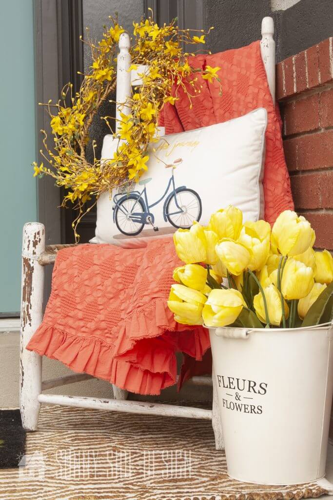 Front porch Spring Decorating ideas, forsythia wreath styled on a chair with coral throw blanket and pillow, and a bucket of yellow tulips.