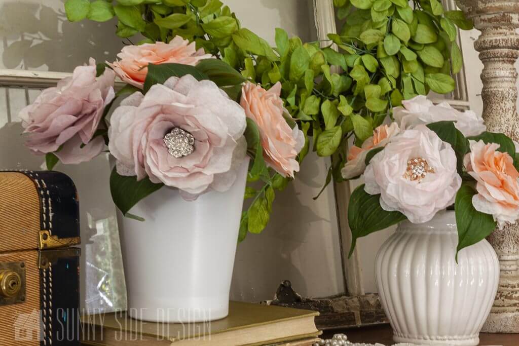 A bouquet of lavender and coral colored coffee filter flowers in rhinestone embellishments in the center for a little extra bling. Flowers arranged in a white vase, styled with a boxwood wreath.
