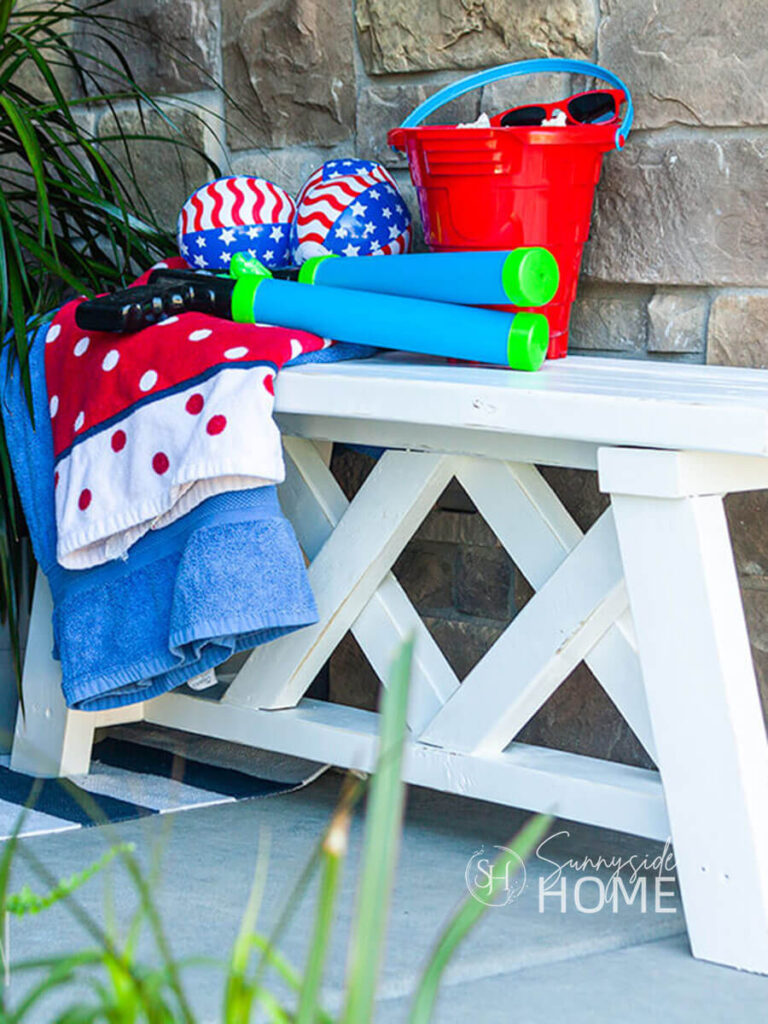 White farmhouse style bench is decorated with red, white and blue beach towels, water guns, patriotic 4th of July beach ball decorations and a red beach pail.