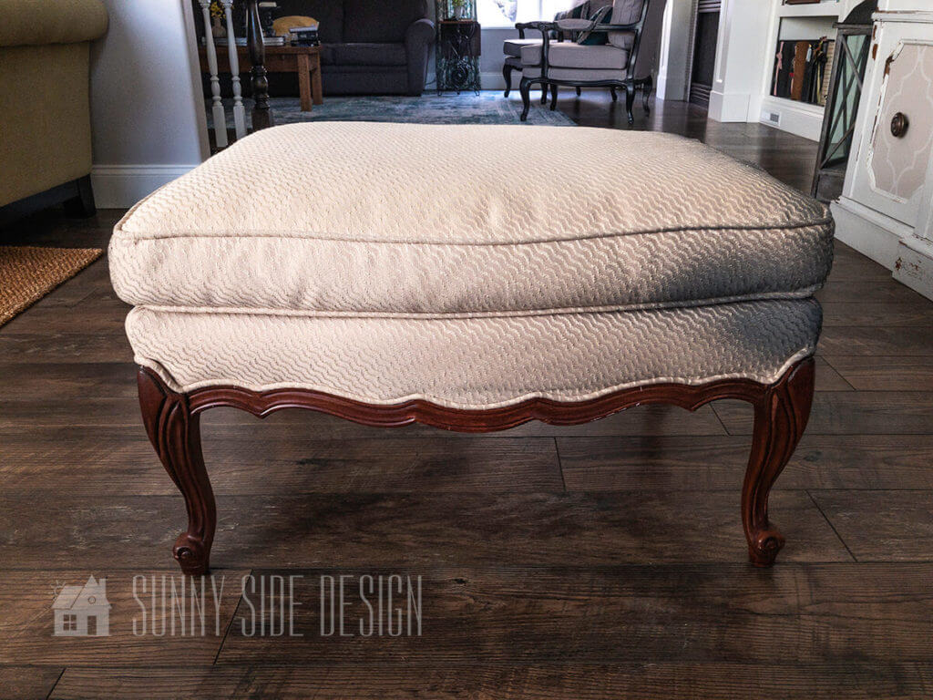 Vintage french style ottoman with cream fabric and walnut toned wood before it is reupholstered.