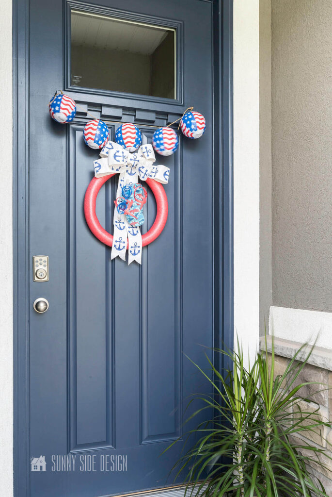 Navy blue front door with 4th of July decorations, a patriotic beach ball bunting placed above a red pool noodle wreath embellished with blue and white nautical ribbon bow and blue swim goggles and patriotic flip flops.