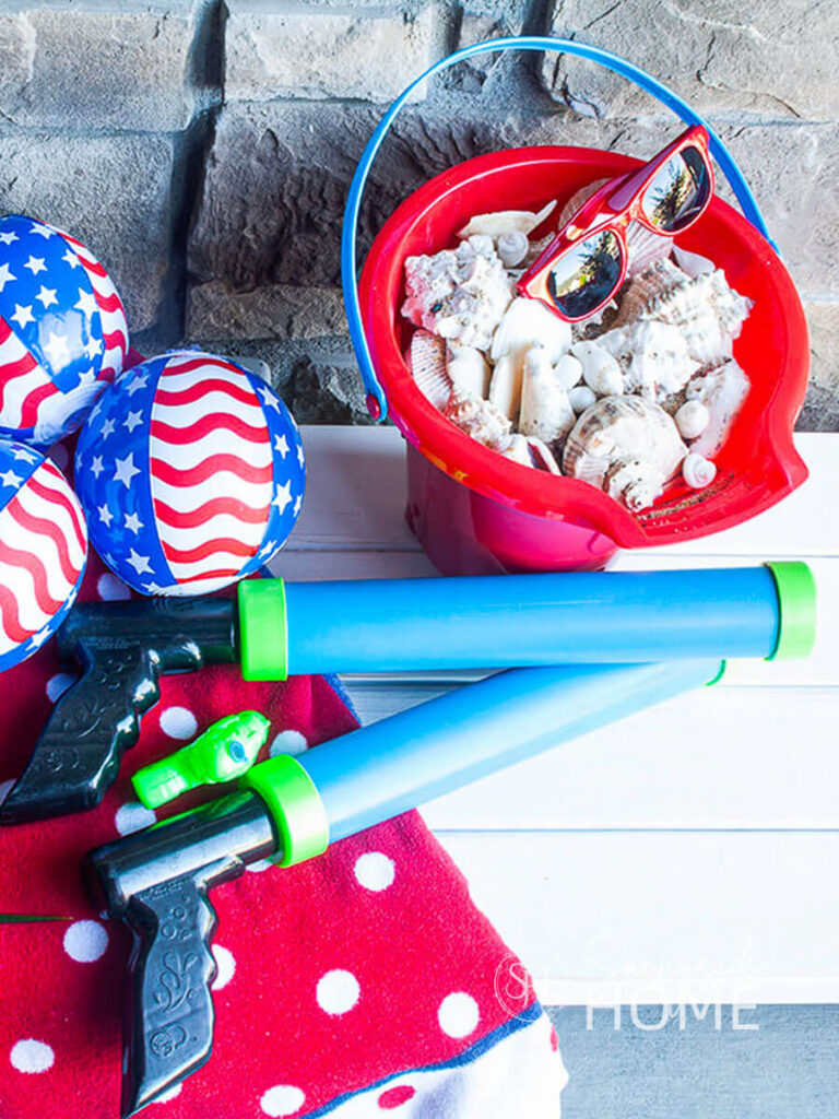 White farmhouse style bench is decorated with red, white and blue beach towels, water guns, patriotic 4th of July beach ball decorations and a red beach pail filled with seashells and sun glasses.