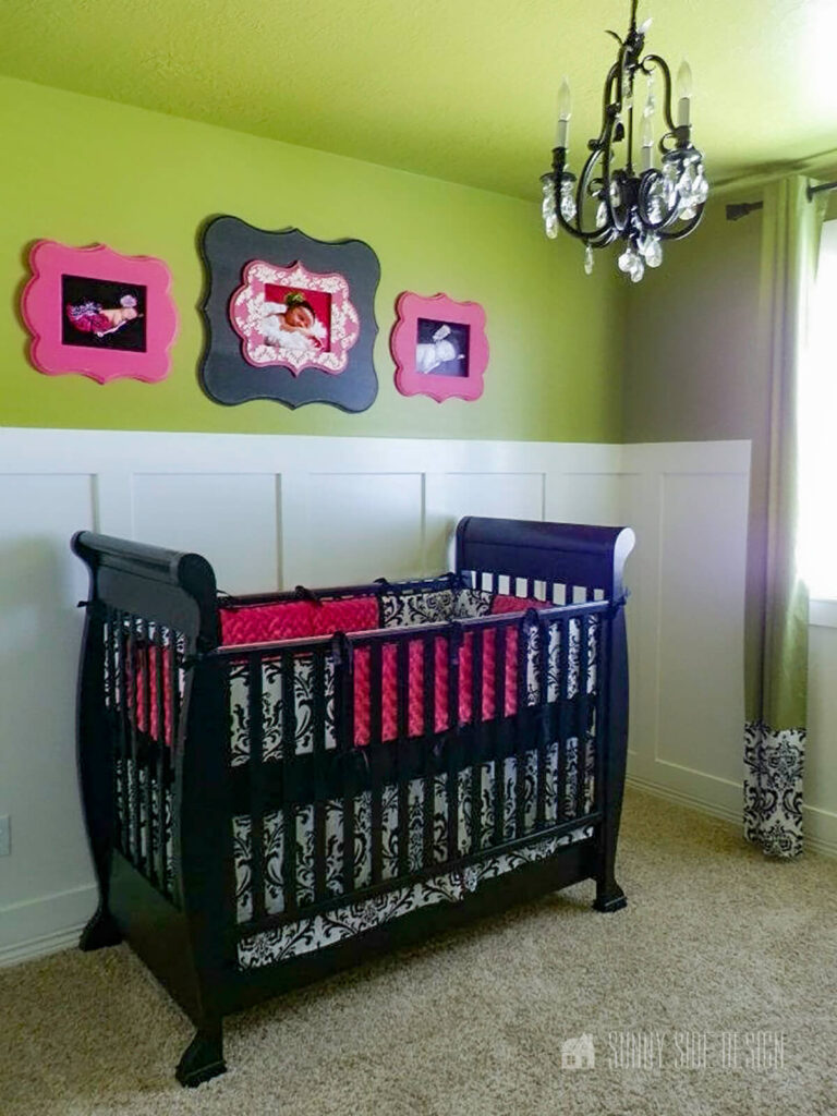 Nursery with white board and batten wall on lower half and avocado green on upper wall and ceiling, black crib and pink and black frames on wall.