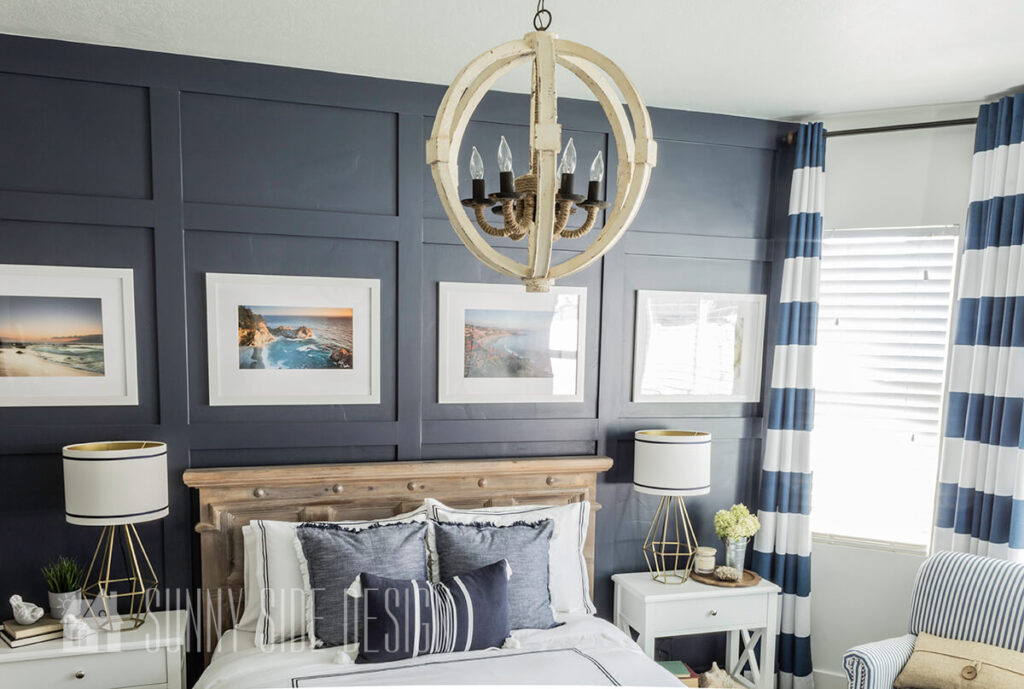 Master bedroom with a navy board and batten wall and a coastal orb chandelier, natural wood headboard, white nightstands and blue and white bedding.