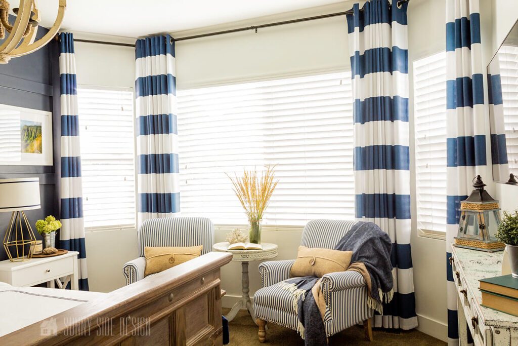 Navy blue and white striped curtains hung to the ceiling in a bay window in a master bedroom. 2 blue and white chairs with an accent table.