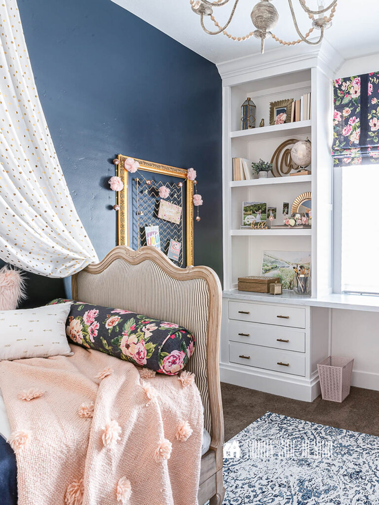 Tween girls bedroom with a French country daybed with Boho pillows, navy blue accent wall, bed crown, built in bookshelves with drawers, built in desk, pink and blue floral roman shades.