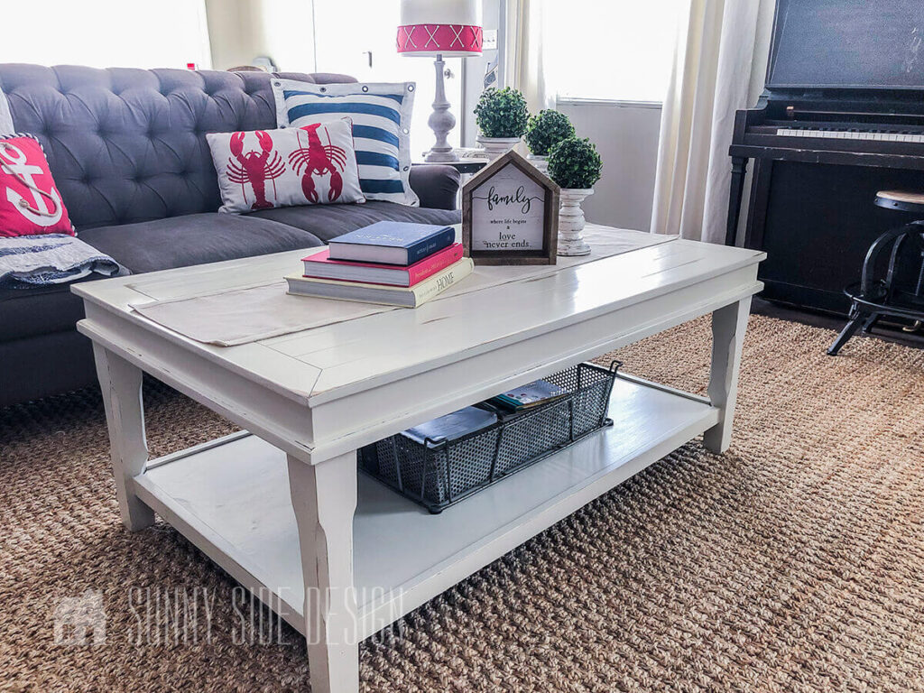White chalk painted coffee table in a living room with a sisal rug, gray tufted sofa, piano and coastal pillows.