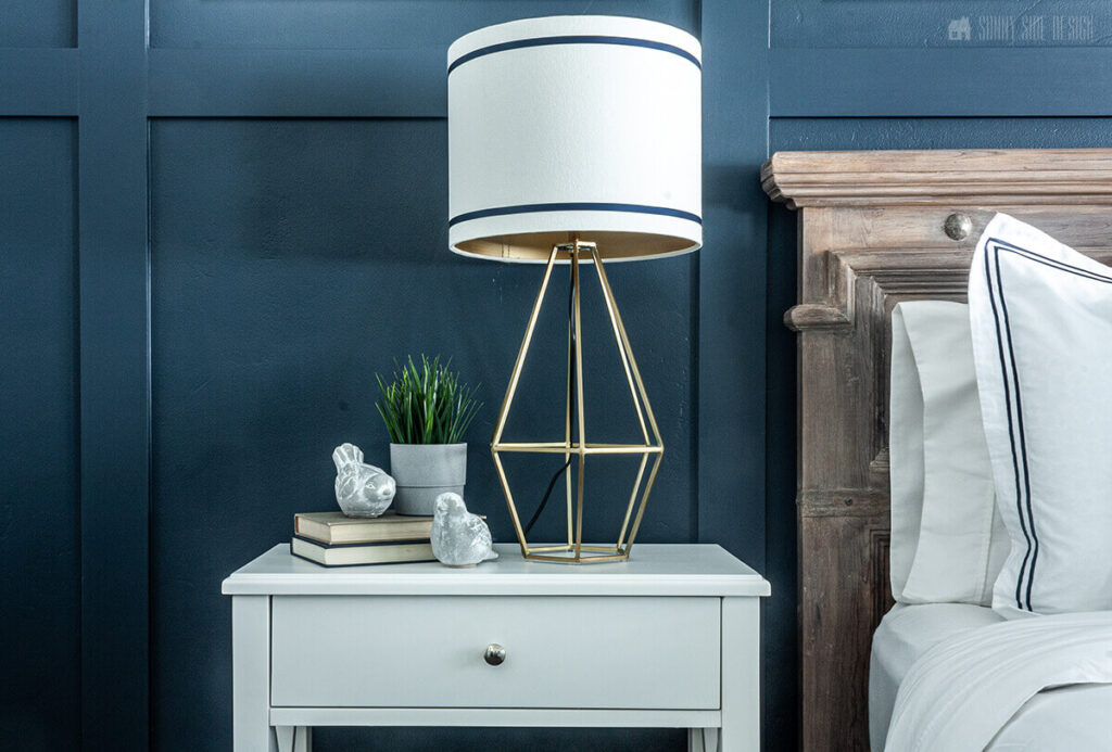 Easily embellish a lampshade with blue grosgrain ribbon. Modern geometric lamp base on a white nightstand with a navy blue board and batten wall.