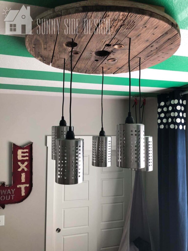 DIY industrial chandelier made with an old electrical spool, Ikea light kit and utensil caddy. The ceiling is painted with wide kelly green strips, wall painted gray with white trim moulding.