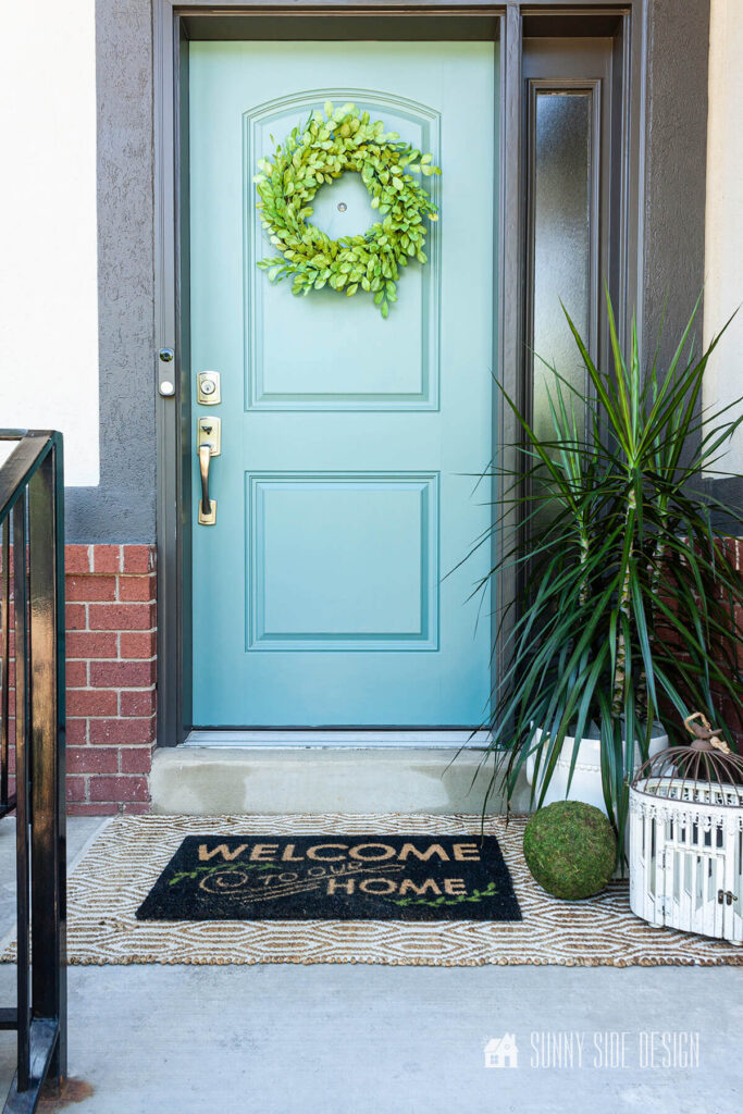 Pretty light blue painted front door with charcoal grey trim, house plant, white birdcage and welcome mat.