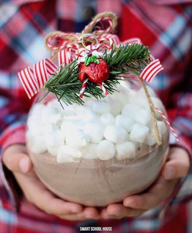 Hands holding a large clear ornament filled with hot cocoa and marshmallows. Embellished with bow and greenery.