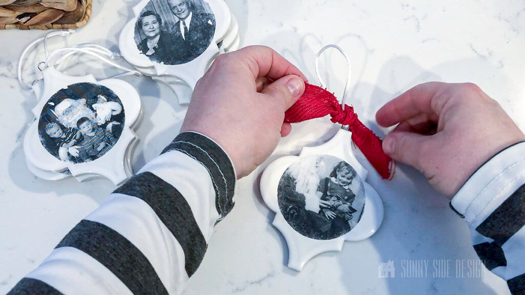 Woman's hands tieing ribbon to the personalized Christmas ornament. Black and white imate of little girl sitting on Santa's lap on a white arebesque ceramic tile
