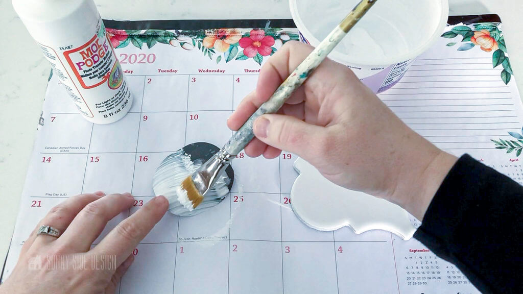 Woman'Woman's hand holding a paint brush applying photo transfer medium to a white arebesque ceramic tile making a personalized Christmas ornament.