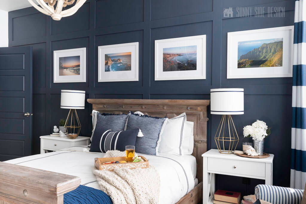 Navy blue board and batten wall a great DIY for beginners, with coatal photography in white frames and matts o the wall. Raw wood bed with white and navy linens, white night stands with geometric gold lamps with white shades accented with navy blue ribbon.
