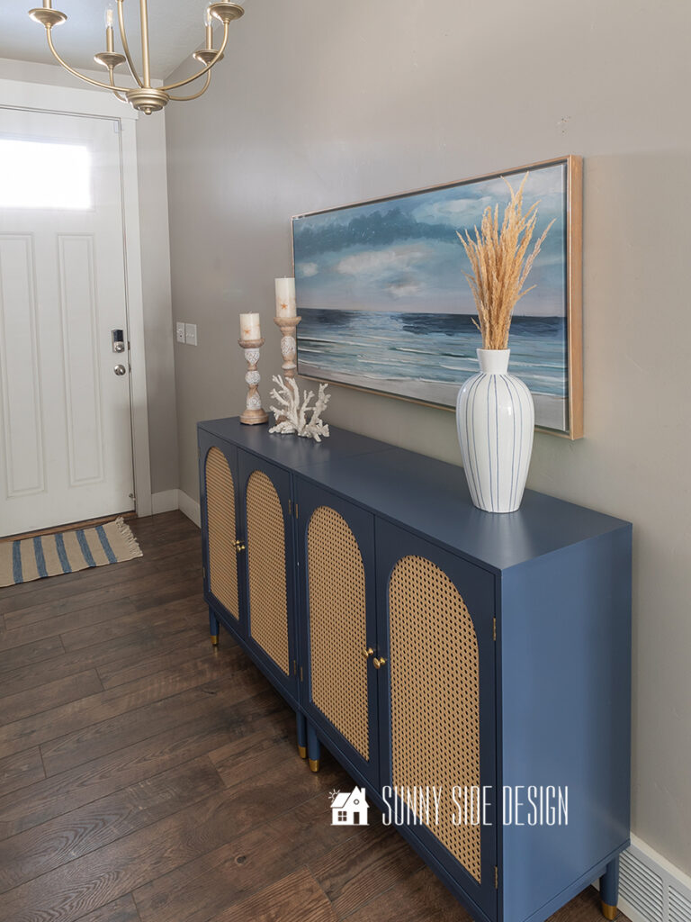 Home tour - entryway with navy blue and cane console table decorated with blue and white striped vase with natural grasses, coastal candlesticks and coral with a coastal painting hung on the wall and a brushed gold chandelier.