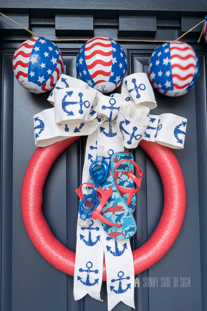 Playful and patriotic Summer Wreath for Front Door made with a red pool noodle, flip flops, swim goggles, and nautical ribbon on a navy blue door with mini patriotic beach ball garland.