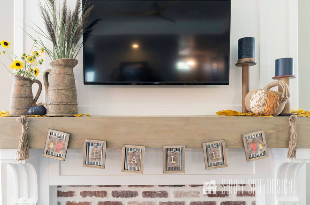 Free Printable Fall Home Decor Bunting hangin from the mantle. The mantle is decorated with to pitchers fillsed with sunflowers and grasses, as well as 2 candlesticks with navy blue taper candles. There is also a textured pumpkin in front of the candlesticks.