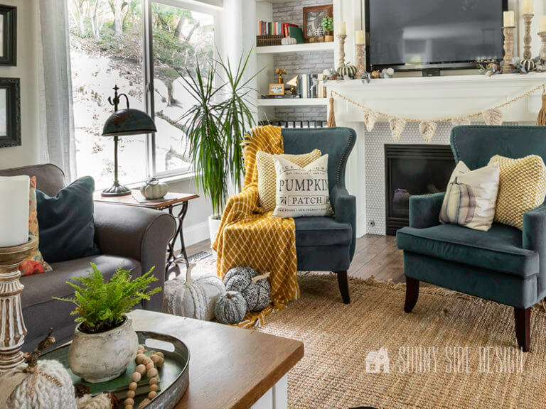 Cheap Fall Decor ideas, 2 peacock blue wingback chairs infront of firepace. Once chair draped with a mustard throw blanket and fall pillows. 3 cozy pumpkins rest on the edge of the throw blanket at the let of the chair.