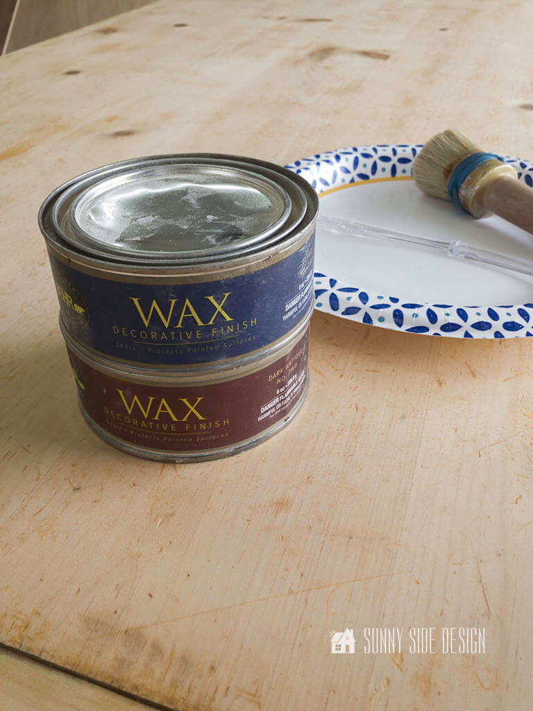 Clear and dark wax cans setting on table top with a waxing brush and paper plate.