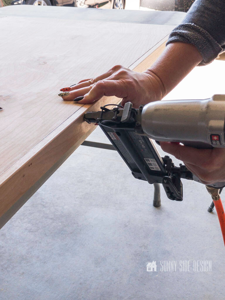 Woman's hand holding pneumatic brad nailer, securing wood trim to the plywood edge of laundry room folding table.