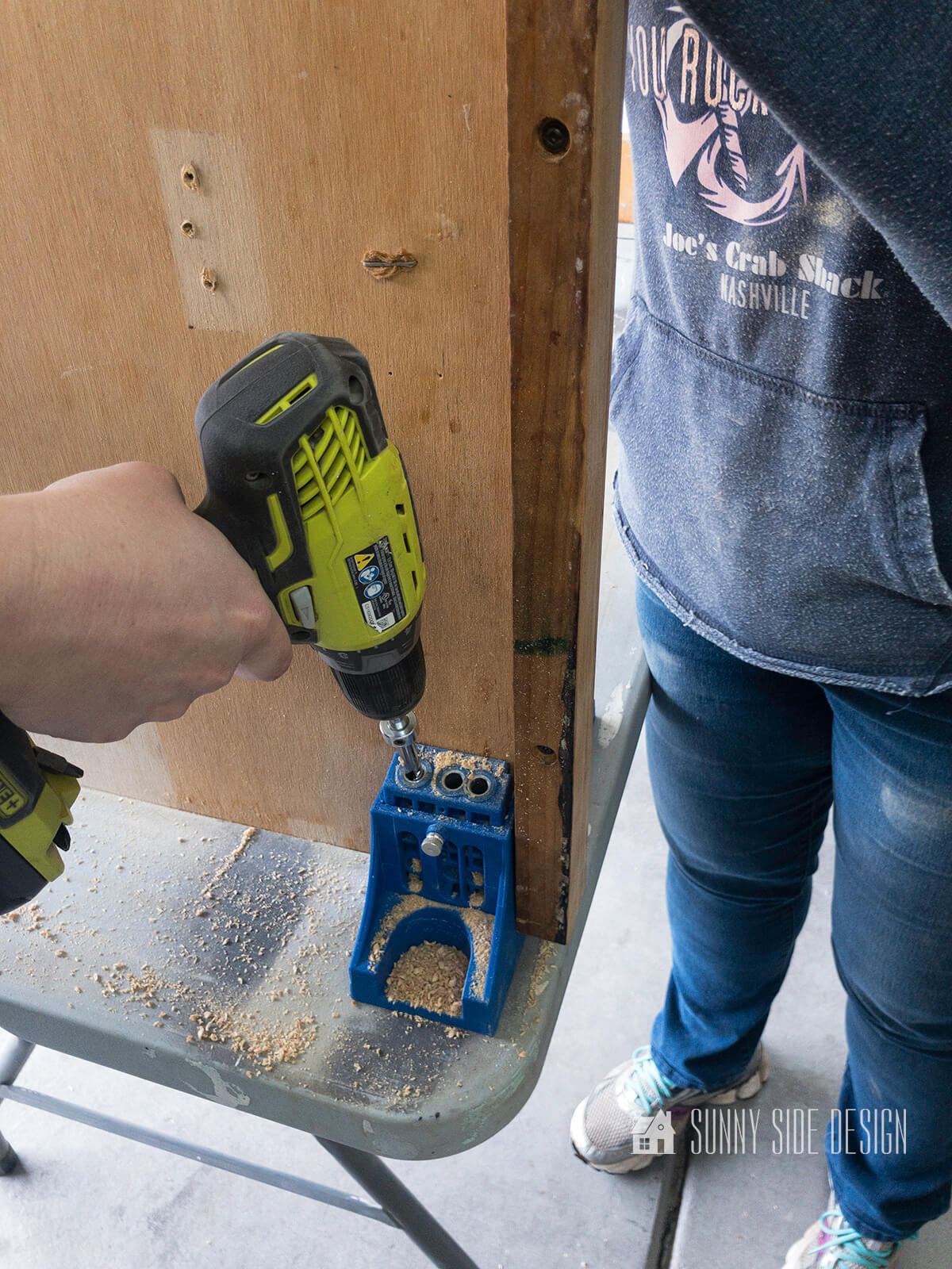 Pocket hole jig is secured to folding table top and woman's hand holds drill, drilling holes for pocket screws.