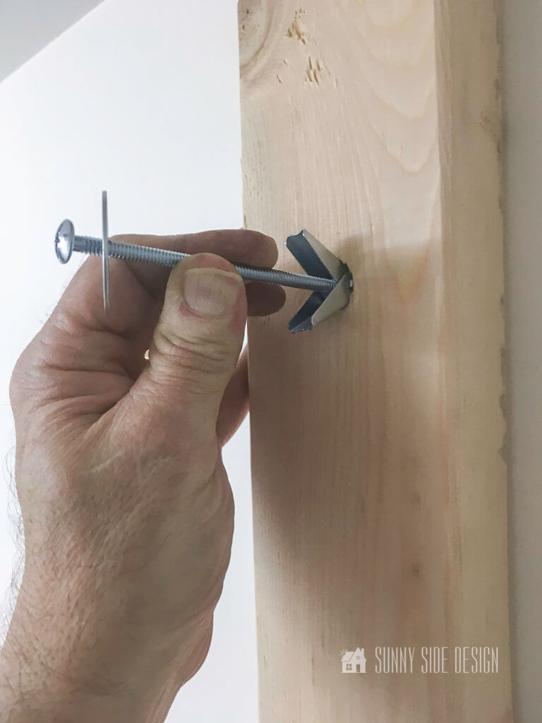 Mans hand plainging a molly bolt through a hold in lumber into the wall.