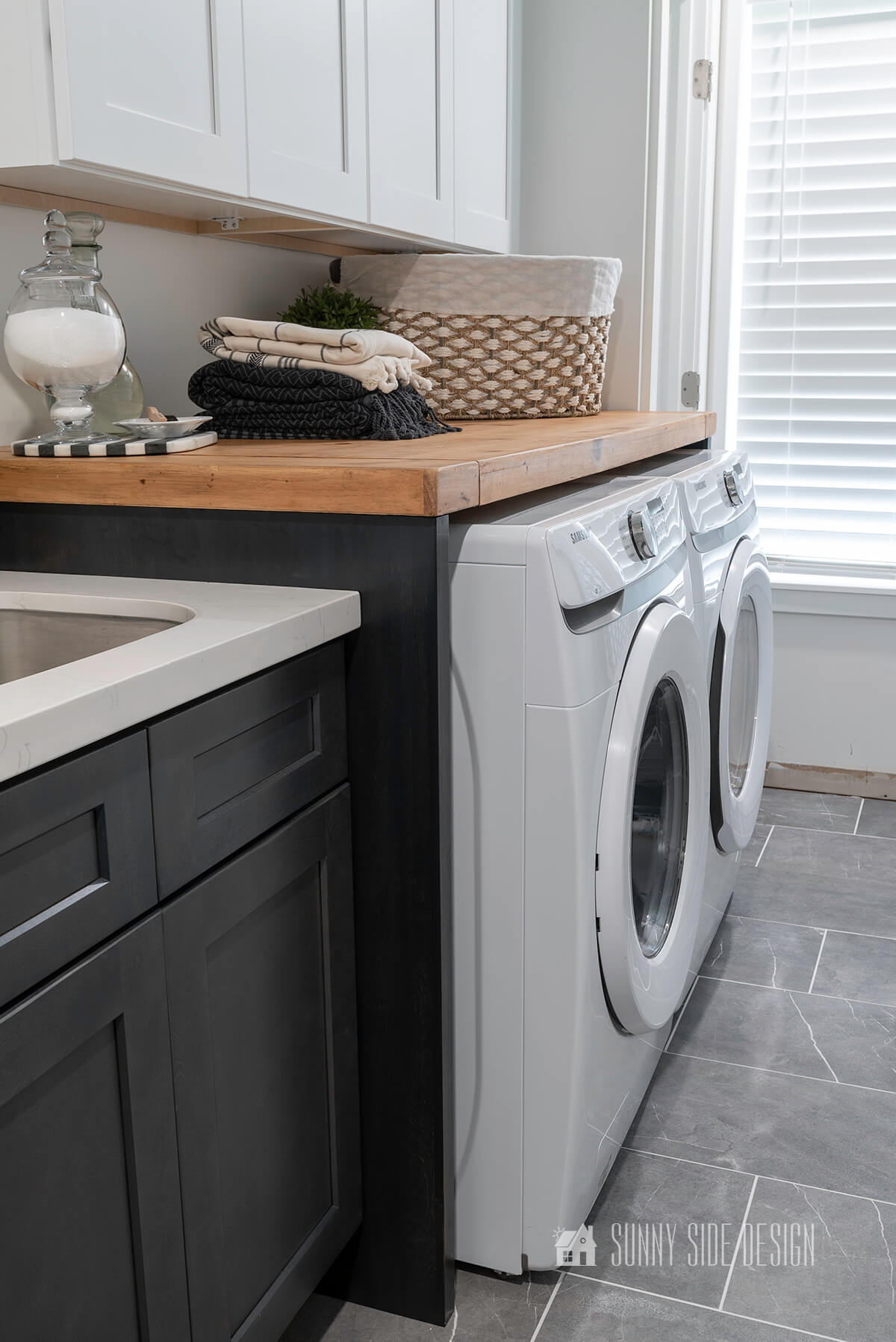 Laundry room with black and white cabinets, quartz countertop and stainless steel sink. White laundry appliances with a natural wood folding table over appliances. Topped with a jute laundry basket and folded towels.