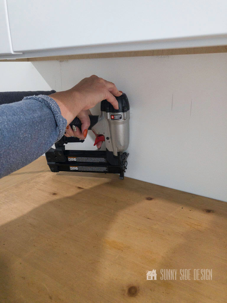 Woman's hand hold pneumatic brad nailer, inserting brads along the back of the laundry room fold table into the cleat on the wall.