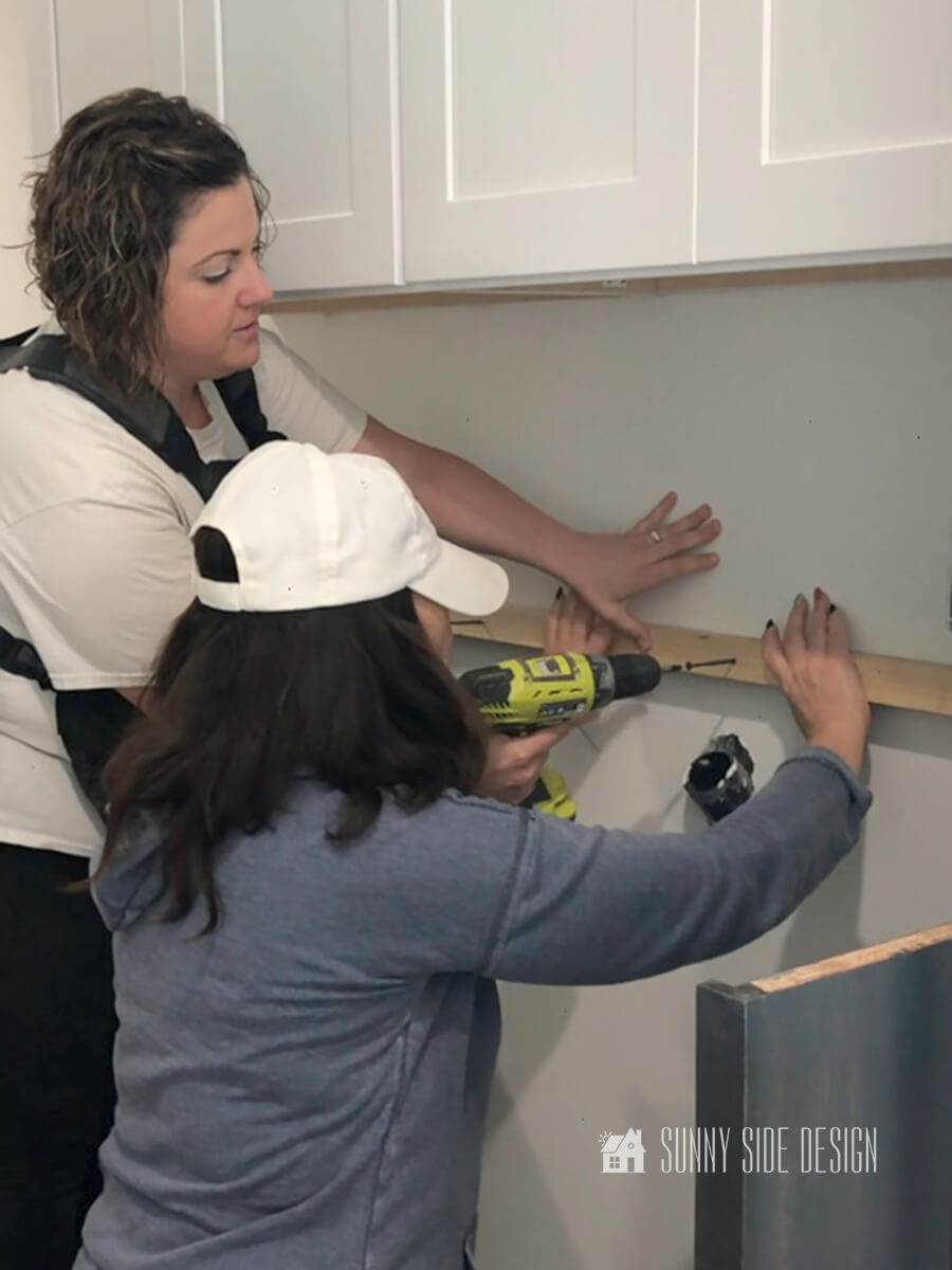 One woman is holding cleat and another woman is inserting a screw through the cleat into the stud in the wall in the laundry room.