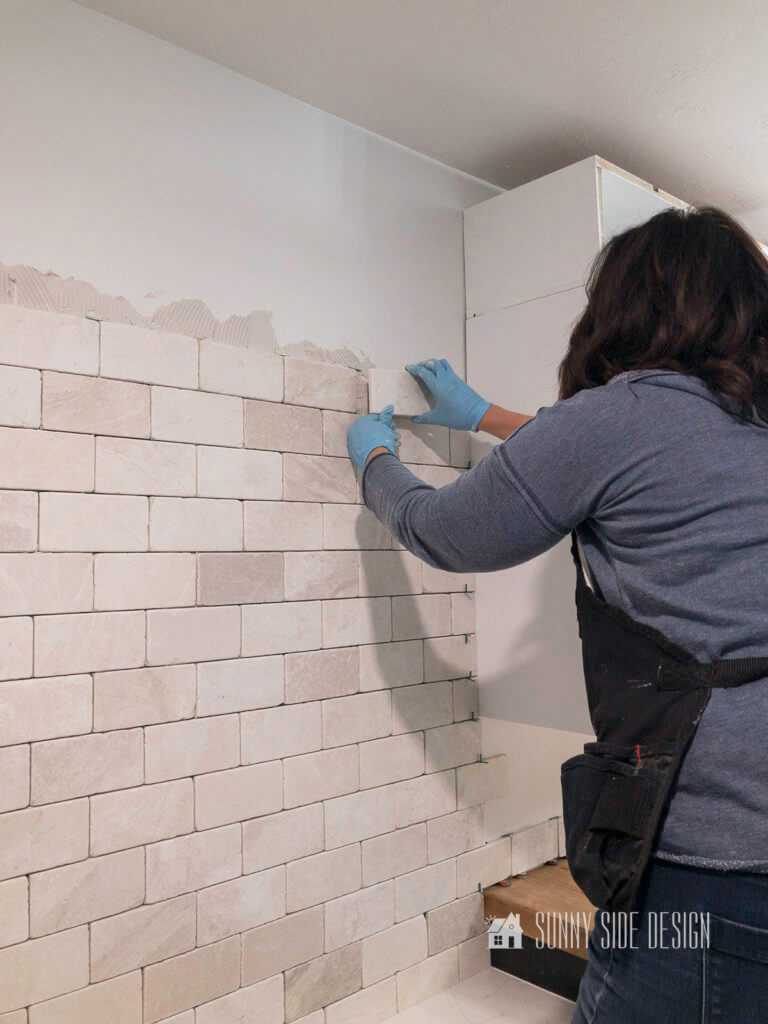 Woman wearing a blue sweatshirt and work apron placing tile onto the wall while install a tile backsplash.