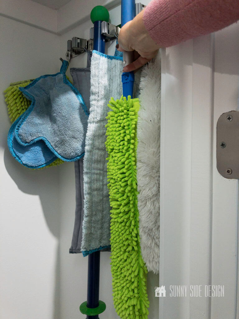Womans hand returning cleaning tool to the hanging storage in the broom closet. Also hanging on the organizer is dusting mits, dust mop, mop.