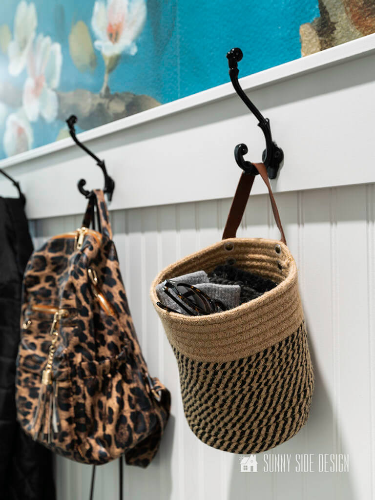 Laundry room Organization ideas by adding hooks for a mudroom along one wall. Black hooks on a bead board wall with a blue floral wallpaper. Hooks are holding a bag for gloves, hats and glasses and a backpack.