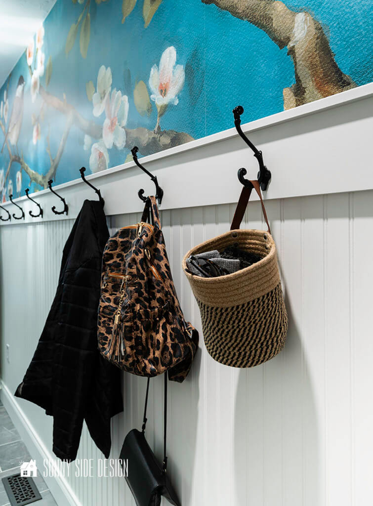 Laundry room Organization ideas by adding hooks for a mudroom along one wall. Black hooks on a bead board wall with a blue floral wallpaper. Hooks are holding a bag for gloves, hats and glasses and a backpack.