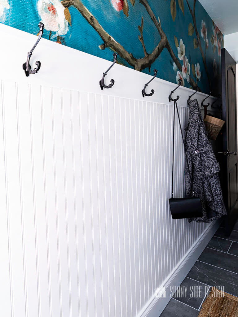Mudroom wall with bead board, blue floral wallpaper and black hooks, purse and coat.
