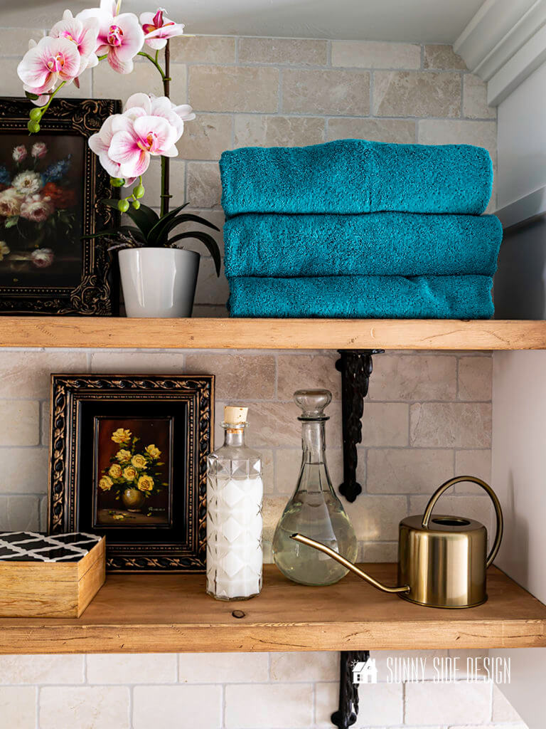 Laundry room makeover, marble tiled wall with natural wood whelves and black iron brackets, accessorized with stacked blue towels, pink orchid, vintage glass containers filled with detergent and fabric softner and a wood black and white box.