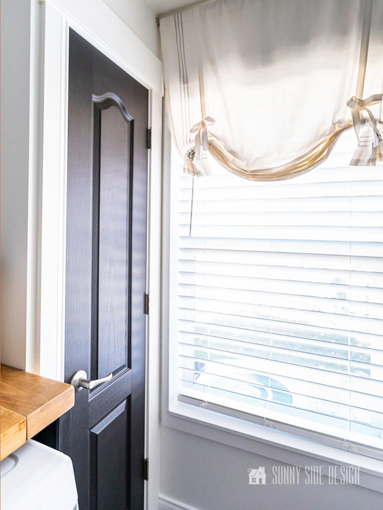 Black door to broom closet in laundry room. Window with a white and grey striped balloon shade.