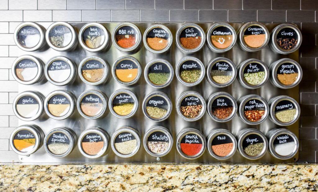 Round metal and glass spice jars are label and store with magnets on a metal sheet mounted on the backsplash.
