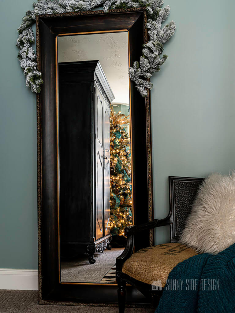Black floor mirror topped with a flocked garland leaning against a slate blue wall. Next to mirror is a black cane chair with a fur pillow and peacock blue throw blanket. In the reflection on the mirror you can see the lights from the Christmas tree.