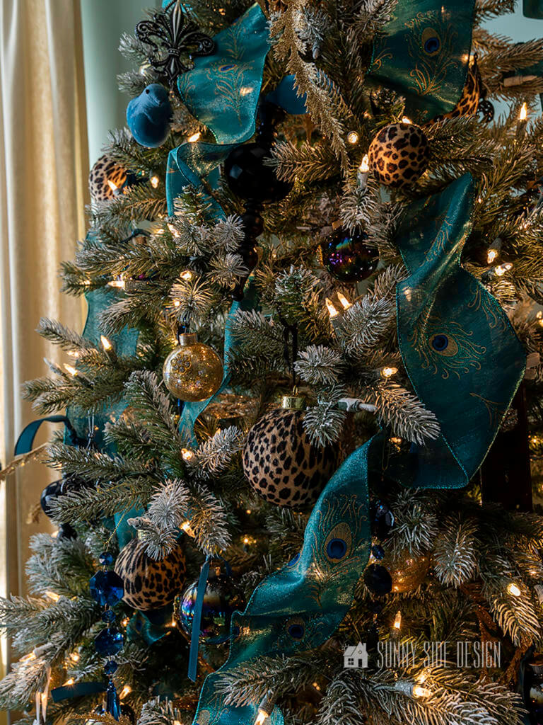 Flocked Christmas tree with cheetah ball ornaments, gold glitter ball ornaments, blue jewels, blue birds, black glitter ball ornaments, gold leaf picks and peacock blue ribbon.