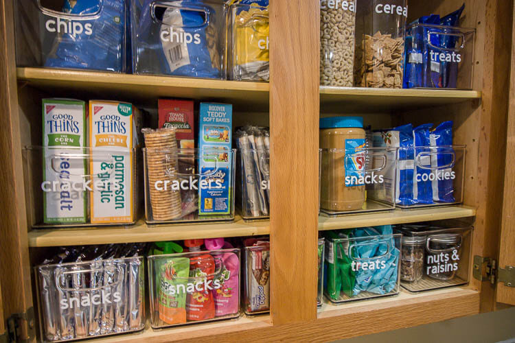 Pantry Organization Ideas - Clear rectangle containers labeled with white vinyl lettering store packaged food in a cupboard. Organized by categories, like crackers, chips, snacks, cookies etc.