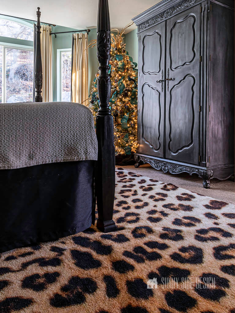 Decorate bedroom for Christmas with a full size Christmas tree with twinkling lights. 4 poster black bed with a black dust ruffle, beige quilted coverlet, black armoire and a cheeta print rug.