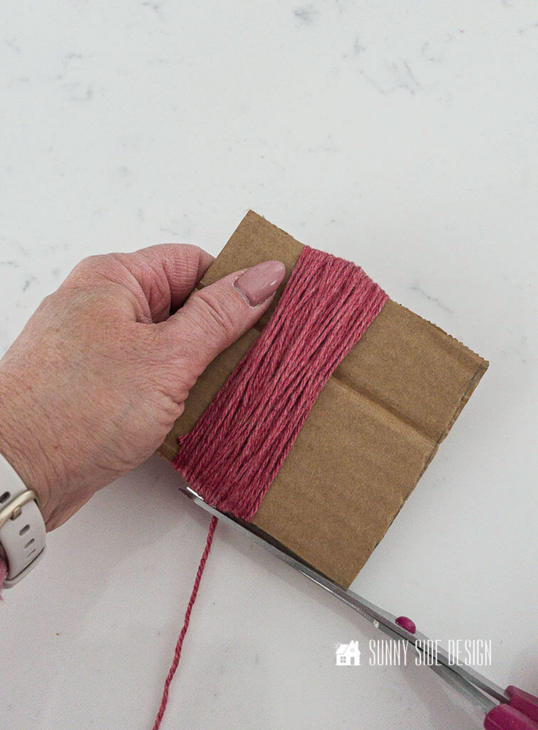 Woman's hand with wrapped dusty pink yarn wrapped around a square of cardboard and scissors are trimming the yarn.