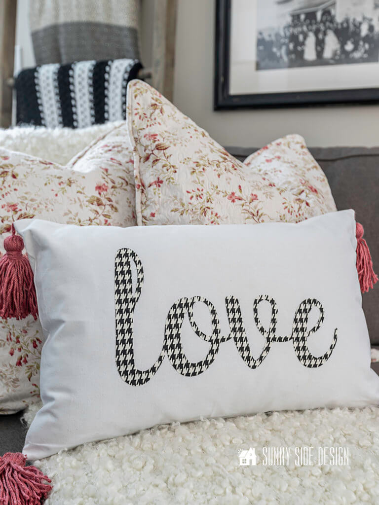 Pillow cover with black and white houndstooth "love" and dusty rose tassels, sitting on sofa with floral pillows.